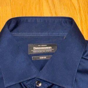 Mörkblå skjorta Dressmann slim fit - Snygg mörkblå skjorta från Dressmann i slim fit-modell. Skjortan har klassisk krage, svarta knappar och är tillverkad i bomull med stretch för extra komfort. Perfekt för dig som gillar stilrena och enkla plagg med bra passform. Storlek L. Säljes pågrund av för stor aldrig använd 