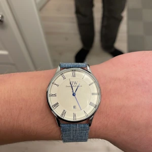 Daniel Wellington klocka med blått tygband - Snygg klocka från Daniel Wellington med rund silverfärgad boett och vit urtavla med romerska siffror. Blå visare och datumvisning. Bandet är i blått tyg med silverfärgat spänne, vilket ger en chill och modern vibe. Perfekt för dig som gillar stilrena accessoarer.