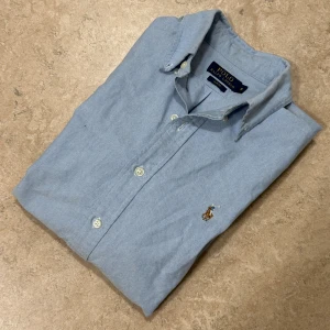 Blå skjorta Polo Ralph Lauren slim fit - Snygg ljusblå skjorta från Polo Ralph Lauren i slim fit-modell. Klassisk krage, knappar framtill och broderad logga på bröstet. Tillverkad i mjuk bomull och har långa ärmar. Perfekt för dig som gillar stilrena och tidlösa plagg.