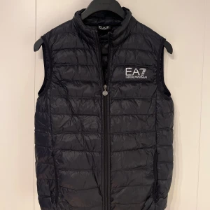 Svart dunväst från EA7 Emporio Armani - Svart quiltad dunväst från EA7 Emporio Armani med silverfärgat logotryck på bröstet. Västen har hög krage, dragkedja framtill och två sidofickor. Materialet är glansigt och lätt, perfekt för lager-på-lager. Snygg och sportig look med stilren design.