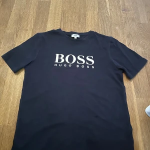 Mörkblå t-shirt från Hugo Boss XS - Snygg mörkblå t-shirt från Hugo Boss i storlek XS med klassisk vit logotyp på bröstet. T-shirten har rund hals och korta ärmar, tillverkad i mjuk bomull som känns skön mot huden. Perfekt för dig som gillar stilrena och enkla plagg med premiumkänsla.