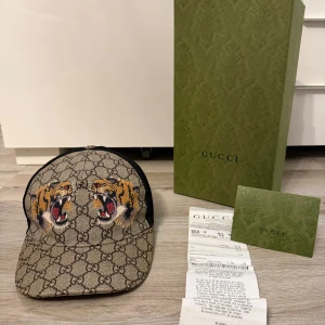 Gucci keps med tiger - Snygg Gucci keps med klassiskt GG-mönster i beige och brunt, prydd med två broderade tigrar framtill. Justerbar rem med gröna och röda ränder bak. Mesh på sidorna för extra ventilation. Lyxig och unik accessoar som sticker ut. Box och kvitto följer med:) ÄKTA. Vid frågor eller funderingar så är det bara att höra av sig!!