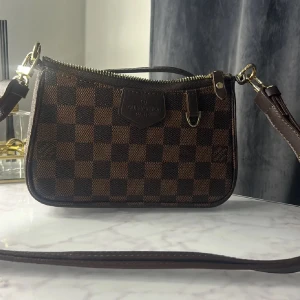  brun axelväska rutig - Snygg axelväska  klassiskt brunt rutigt mönster med guldfärgade detaljer och dragkedja. Väskan har justerbar rem och logotyp framtill. Perfekt storlek för det viktigaste och ger en lyxig touch till din outfit.har gått sönder lite i handtagen men går att fortfarande än vända 