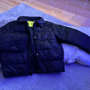 Svart pufferjacka från JUXX - Svart pufferjacka från JUXX med klassisk krage och quiltad design. Jackan har dragkedja och tryckknappar framtill, samt en gul insida med stor logotyp. Perfekt för kalla dagar och har en clean, modern look.