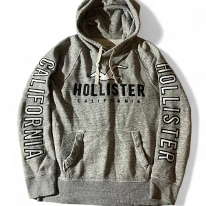 Grå Hollister hoodie med tryck XS/S - Snygg grå hoodie från Hollister i storlek XS med stor logga och texten CALIFORNIA på bröstet och längs båda ärmarna. Tröjan har huva med snörning och en stor magficka framtill. Mjuk insida och klassisk streetstil.