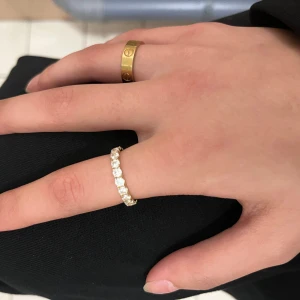 Guldring med vita stenar - Snygg guldring med gnistrande vita stenar runt hela ringen. Ringen har en klassisk och elegant design med rund form och stenar som ger extra glans. Perfekt för dig som gillar bling och vill ha något som sticker ut på handen.