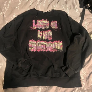 Svart sweatshirt Lost in the Moment - Svart sweatshirt med färgglatt tryck 'Lost in the Moment' i lila, rosa och grönt. Tröjan har rund hals, ribbade muddar och lång ärm. Perfekt för dig som gillar streetwear och grafiska prints.