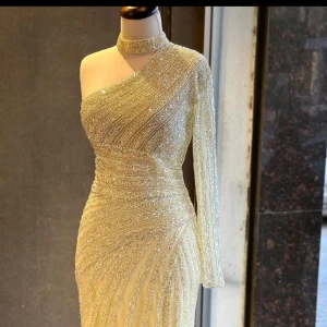 Champagnefärgad glittrig långklänning - Supersnygg långklänning i champagnefärg med glittriga detaljer och ett elegant mönster. Klänningen har en one shoulder-design med lång ärm och hög halsringning. Materialet är skimrande och ger en lyxig känsla, perfekt för att sticka ut på bal eller fest.