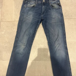 Replay Anbass blå jeans herr - Snygga Replay Anbass jeans i klassisk blå tvätt med coola slitningar och detaljerade sömmar. Modellen har fem fickor, normal passform och är tillverkad i stretchigt jeanstyg för extra komfort. Perfekta för dig som gillar en stilren och modern look. Längd 32