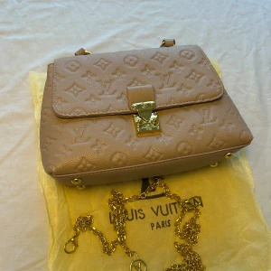 Beige Louis Vuitton handväska i skinn - Snygg beige handväska med klassiskt monogrammönster i skinn. Väskan har guldfärgade metalldetaljer, en elegant kedja och ett stilrent lås framtill. Perfekt storlek för det viktigaste och en lyxig touch till din outfit.