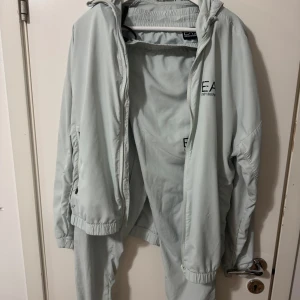 Ljusgrå vindjacka från EA7 Emporio Armani - Snygg ljusgrå vindjacka från EA7 Emporio Armani med huva och dragkedja framtill. Jackan har två sidofickor med dragkedja och elastiska muddar vid ärmslut och nederkant. Perfekt för en sportig och clean look.