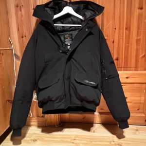 Svart dunjacka från Canada Goose XL - Canada Goose dunjacka i svart med huva och klassisk logga på ärmen. Jackan har stora fickor med lock, dragkedja och knappar framtill samt ribbade muddar vid ärmslut. Perfekt för kalla dagar och har en clean, urban look.