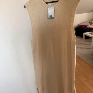 Beige stickad ärmlös klänning H&M - Stickad beige klänning från H&M i storlek M. Modellen är ärmlös med rund hals och har en rak, lång siluett. Materialet består av bomull, varav 20% är återvunnen. Perfekt för dig som gillar enkel och clean stil.