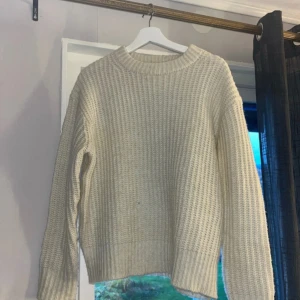 Beige stickad tröja med rund hals - Säljer en beige stickad tröja med grov struktur och rund hals. Tröjan har långa ärmar och breda ribbade muddar vid ärmslut och nederkant. Perfekt för att hålla sig varm under kyliga dagar. Enkel och stilren design som funkar till många olika outfits.