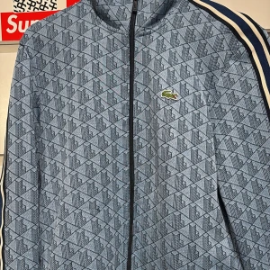 Blå mönstrad kofta från Lacoste - Snygg blå kofta från Lacoste med geometriskt mönster och klassisk krokodillogga på bröstet. Jackan har svarta och vita ränder längs axlarna, dragkedja framtill och regular fit. Perfekt för dig som gillar sportig och stilren streetwear.