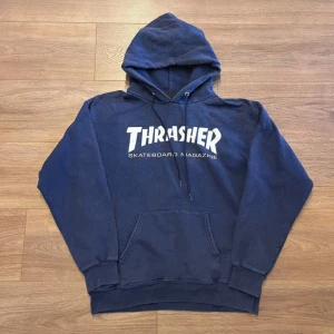 Mörkblå Thrasher hoodie med huva - Säljer en mörkblå hoodie från Thrasher med klassisk logga i vitt på bröstet. Tröjan har huva med snörning och en stor magficka. Perfekt för dig som gillar skate och streetwear. Passar dig som vill ha en chill och snygg look.