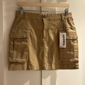 Beige cargo-kjol från Kappahl - Snygg beige cargo-kjol från Kappahl med stora fickor på sidorna och knappstängning fram. Kjolen har bälteshällor och är tillverkad i mjuk bomull, perfekt för en chill och avslappnad stil. Passformen är normal och kjolen är kort.