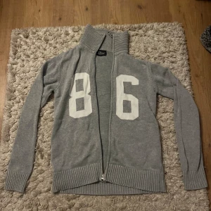 Grå kofta Peak Performance med dragkedja - Snygg grå stickad kofta från Peak Performance i storlek S. Framsidan har vita siffror 86 och koftan har en ribbad hög krage samt hel dragkedja. Perfekt för dig som gillar sportig och chill stil.