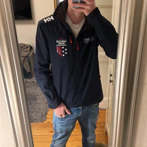 Helly Hansen American Magic vindjacka - Snygg marinblå vindjacka från Helly Hansen med American Magic-tryck och coola detaljer som vita och röda stjärnor och ränder. Jackan har dragkedja, hög krage och röda zip-pullers. Perfekt för dig som gillar sportig stil och vill sticka ut.