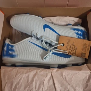 Nike Mercurial blå fotbollsskor - Nike Mercurial fotbollsskor i ljusblått med starkt blå detaljer och Swoosh-logga på sidan. Skorna har snörning, platt sula med dobbar och är tillverkade i syntetmaterial. Perfekt för dig som vill sticka ut på planen med stilren och sportig look.