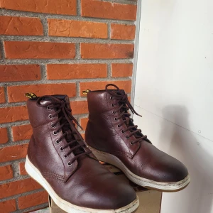 Bruna Dr. Martens boots i skinn - Säljer ett par snygga bruna boots från Dr. Martens i äkta skinn. Modellen har snörning framtill, rund tå och vit platt sula med klassisk gul logotag baktill. Perfekt för dig som gillar robust stil och vill ha något som sticker ut.