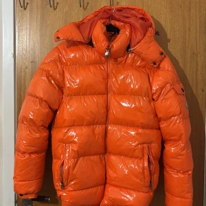 Orange dunjacka från Moncler - Säljer en fet orange dunjacka från Moncler med glansig finish och huva. Jackan har dragkedja framtill, två fickor med dragkedja och Moncler-logga på ärmen. Perfekt för kalla dagar när du vill sticka ut med färg. Riktigt puffig och varm vibe.