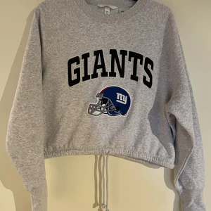 Grå croppad Giants sweatshirt H&M - Grå sweatshirt från H&M med stort GIANTS-tryck och NFL-logga framtill. Croppad modell med snörning i midjan och lång ärm. Mjuk insida och klassisk crewneck. Perfekt för dig som gillar sportig stil och amerikansk fotboll.