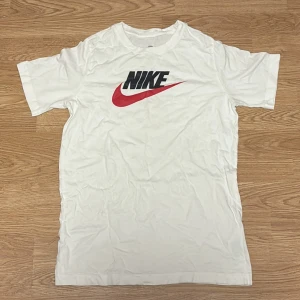 Vit Nike t-shirt med röd logga - Snygg vit t-shirt från Nike med klassisk svart och röd logga på bröstet. Modellen är kortärmad och har en rund hals. Hör av er vid intresse!