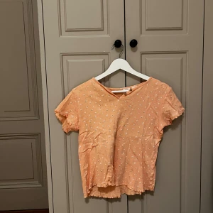 Persikofärgad blommig topp från Zara - Supersöt persikofärgad topp från Zara med små vita och rosa blommor över hela. Toppen har v-ringning med två knappar, korta ärmar med volangkant och är tillverkad i mjuk bomull. Perfekt för dig som gillar gulliga detaljer och somriga färger!