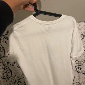 Vit Alpha Industries t-shirt M - Snygg vit t-shirt från Alpha Industries i storlek M. Klassisk rund hals och korta ärmar. Stor svart logga och text på bröstet ger en clean och modern vibe. Perfekt för dig som gillar streetwear och enkel stil. Materialet är mjuk bomull som känns skönt mot huden.