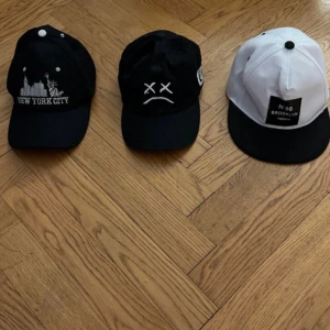 Tre kepsar: Brooklyn, NYC och smiley - Säljer tre kepsar för 120kr! En keps för 45 kr!  en svart med broderad New York City och Frihetsgudinnan, en svart med vit smiley och en vit/svart keps från District Twenty med Brooklyn-tryck. Alla har böjd skärm och justerbar baksida. Perfekta accessoarer för en urban streetstyle.