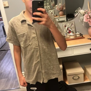 Beige frottéskjorta från Kappahl - Beige skjorta från Kappahl i mjuk frotté med kort ärm, klassisk krage och bröstficka. Skjortan har knäppning framtill och en avslappnad passform som känns chill och trendig.