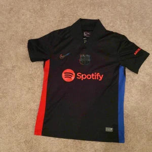 FC Barcelona svart Nike fotbollströja S - Säljer en FC Barcelona matchtröja från Nike i svart med röda och blå sidopaneler. Tröjan har korta ärmar, krage och tryck med Spotify och UNHCR-logga. Materialet är Dri-FIT polyester som andas och håller dig torr. Perfekt för dig som älskar Barca! OBS Nike loggan är lite lös