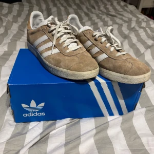 Adidas Gazelle beige mocka sneakers - NYTT SKICK HELT OANDVÄNDA!                    Säljer ett par Adidas Gazelle sneakers i beige mocka med vita detaljer och klassiska tre ränder på sidan. Skorna har rund tå, platt sula och snörning. Ikonisk retrostil med vit Adidas-logga på hälen och tungan. Perfekt för dig som gillar clean och stilren look.