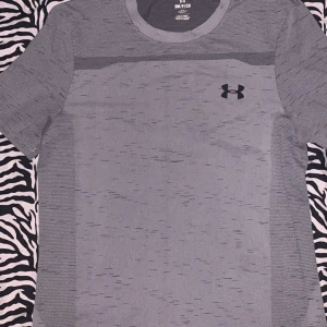 Grå Under Armour t-shirt, slim fit - Snygg grå t-shirt från Under Armour med diskret mönster och svart logga på bröstet. Slim fit-modell med rund hals och korta ärmar, perfekt för träning. Materialet är mjukt och andas, vilket gör den skön att ha på sig under aktiva dagar.