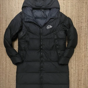 Svart lång pufferjacka från Nike - Svart lång pufferjacka från Nike med huva och två sidofickor med dragkedja. Jackan har ett stilrent Nike-logga på bröstet och är fodrad för extra värme. Perfekt för kalla dagar och har en snygg, enkel design som funkar till streetwear.