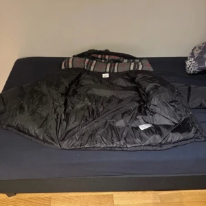 Svart dunjacka från burberry - Säljer en svart dunjacka från burberry med huva och dragkedja. Jackan har ett rutigt foder i grått, vitt och rött, samt klassisk puffad design. Perfekt för kalla dagar och har en riktigt skön passform. Jackan är långärmad och har fickor på sidorna.