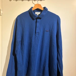 Blå långärmad pikétröja Lacoste M - Klassisk blå långärmad pikétröja från Lacoste med broderad krokodil på bröstet. Tröjan har krage, knappslå och är tillverkad i bomull. Regular fit och perfekt för dig som gillar stilren och sportig look.