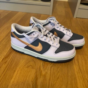 Nike Dunk Low i lila och mörkblå - Nike Dunk Low sneakers med lila och mörkblå paneler, metallic kopparfärgad swoosh och vit broderad Nike-logga på hälen. Skorna har snörning, platt sula och rund tå. Materialet är skinn och syntet, och sulan är svart med bra grepp. Perfekt för dig som gillar unika färgkombinationer.