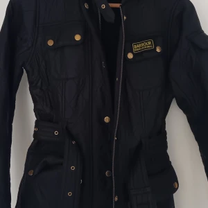 Svart quiltad jacka från Barbour - Svart quiltad jacka från Barbour International med guldiga knappar och bälte i midjan. Jackan har flera fickor med lock, dragkedja och Barbour-logga på bröstet. Snygg, figurnära passform och långärmad design, perfekt för höst och vinter. Ena fickan är lite sönder i sömmen men inget som syns och går säkert att sy för den som kan :) 
