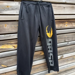 Gasp byxor - Svarta mjukisbyxor från Gasp i storlek S med elastisk midja och snörning. Byxorna har två sidofickor och en stor gul och grå logga längs ena benet. Tillverkade i mjukt syntetmaterial, perfekta för chill eller träning.