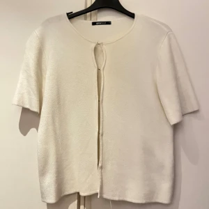 Vit kortärmad kofta/Topp från Gina Tricot - Superfin vit stickad kofta/Topp från Gina Tricot i storlek XS. Den har korta ärmar, rund halsringning och knäpps framtill med små diskreta hakar. 