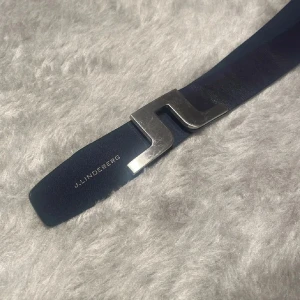 Blått skärp från J.Lindeberg - Snyggt blått bälte från J.Lindeberg med en stilren, silvrig metallspänne i form av märkets logga. Bältet är gjort i skinn och har en modern, minimalistisk look som passar till jeans eller kostymbyxor.