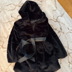 Svart pälsjacka med huva från Amisu - Säljer en svart pälsjacka från Amisu med stor huva och ett snyggt bälte i skinnimitation i midjan. Jackan är fluffig och har långa ärmar, perfekt för kalla dagar. Stilsäker look som verkligen sticker ut.