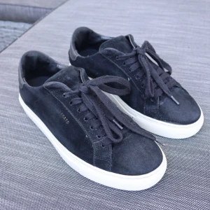 Svarta sneakers från Axel Arigato i mocka - Svarta sneakers från Axel Arigato i mjuk mocka och detaljer i skinn vid hälen. Skorna har rund tå, platt vit sula och klassisk snörning. Snygg minimalistisk design som passar till jeans eller dressade byxor.