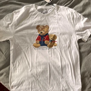 Vit t-shirt med nalletryck från Beverly Hills Polo Club - Snygg vit t-shirt från Beverly Hills Polo Club i 100% bomull. Framsidan har ett gulligt tryck med två nallar, varav en har röd tröja och blå scarf. Regular fit och klassisk rund hals. Perfekt för dig som gillar lekfulla och trendiga motiv.