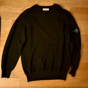 Mörkgrön Stone Island sweatshirt - Mörkgrön långärmad tröja från Stone Island med klassisk patch på vänster ärm. Tröjan har rund hals och ribbade muddar vid ärmslut och nederkant. Perfekt för dig som gillar stilrena och ikoniska streetwear-plagg.