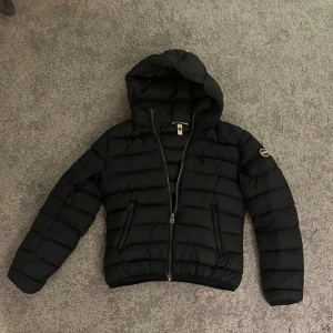 Mörkblå dunjacka med huva från Colmar - Svart dunjacka från Colmar med huva och tvåvägsdragkedja. Jackan har quiltad design, två fickor med dragkedja och Colmar-logga på ärmen. Perfekt för kyliga dagar och riktigt snygg till streetwear-stil.