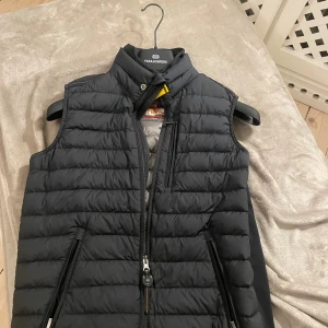 Svart dunväst från Parajumpers - Snygg svart dunväst från Parajumpers med quiltad design och hög krage. Västen har dragkedja framtill, praktiska fickor med dragkedja och en diskret logga på ryggen. Insidan är grå och har märkets autenticitetsmärke. Perfekt för lager-på-lager under kyliga dagar. Den är storlek xs men skulle säga att den passar s också.