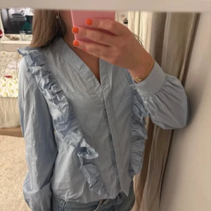 Ljusblå skjorta med volanger - Supersöt ljusblå skjorta med volangdetaljer längs axlar och framstycke. Skjortan har v-ringad hals och långa ärmar med lätt puff vid mudden. Perfekt att styla med jeans för en chill och trendig look.